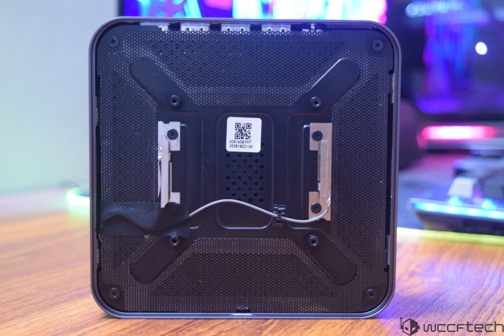 The bottom view of a mini PC, showing a QR code with serial number '2608180010B' and a mounting bracket, placed on a wooden d
